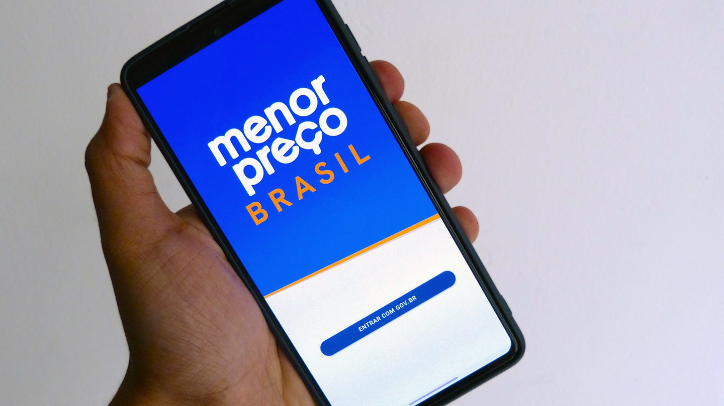 Mão segura celular exibindo tela inicial do Menor Preço Brasil. A tela exibe o logotipo do aplicativo e um botão "Entrar com Gov.br".